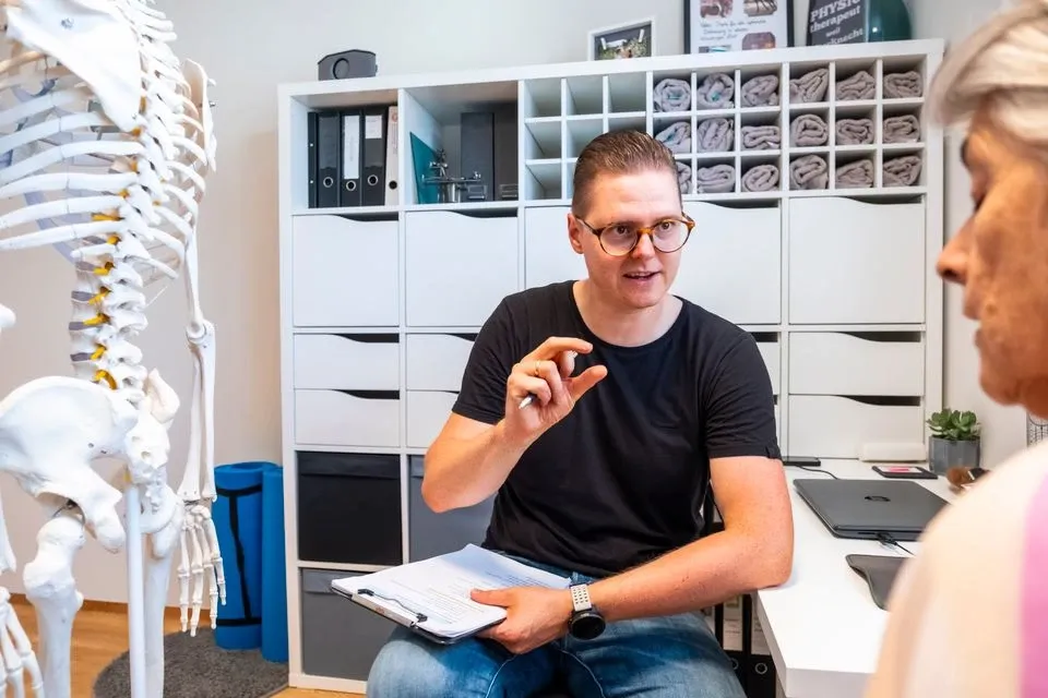Therapeut mit Brille erklärt einer Patientin etwas im Beratungszimmer, im Hintergrund ein Skelettmodell und ein Regal mit Ordnern.