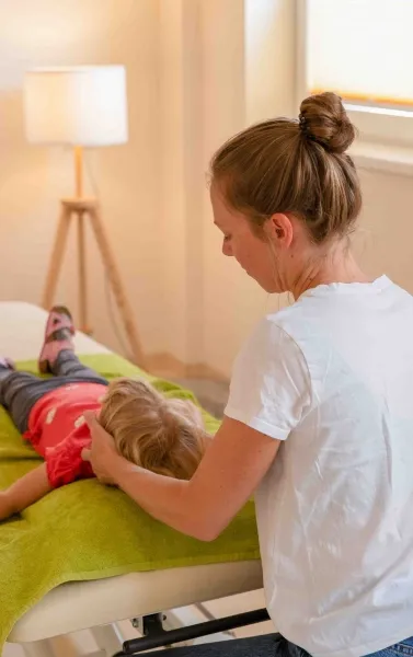 Frau mit zusammengebundenem Haar massiert Kind, das auf einer Liege mit grünem Tuch liegt, im hellen Raum mit Stehlampe