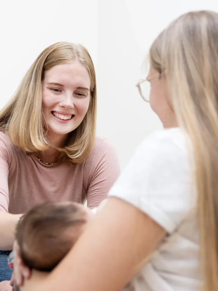 Frau sitzt auf dem Boden mit einem anatomischen Modell des weiblichen Beckens und hält eine Babypuppe, im Hintergrund ein Poster mit einer schematischen Darstellung der Nachgeburt.