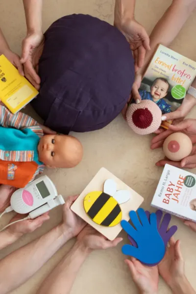 Draufsicht auf einen Kreis aus Händen, die verschiedene Gegenstände rund um Schwangerschaft und Familie in die Mitte halten, darunter eine Babypuppe, ein Stillmodell und Fachbücher.