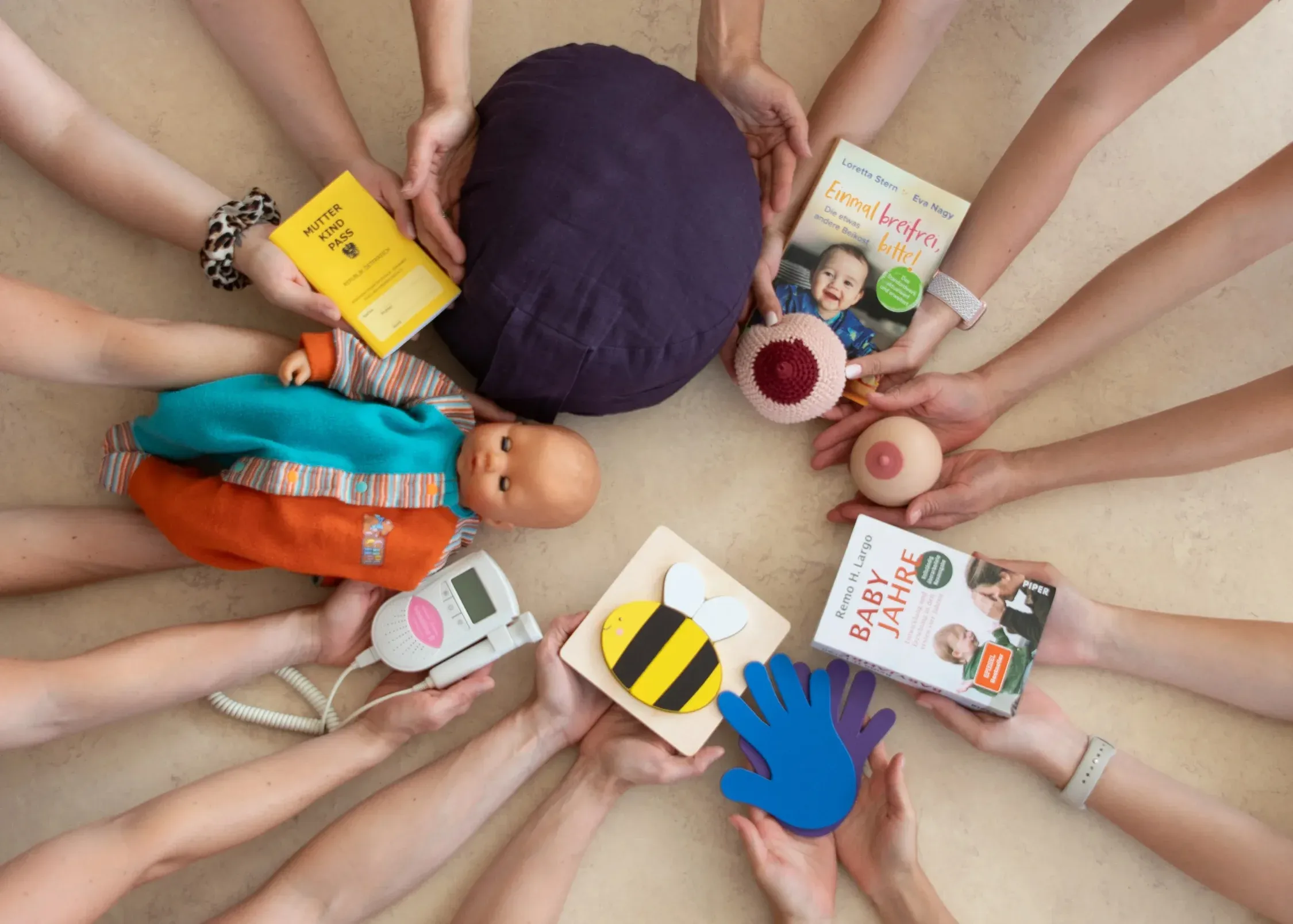 Viele Hände strecken sich von außen im Kreis zur Mitte und halten dabei Gegenstände wie einen Mutter-Kind-Pass, Bücher, ein Stillmodell, ein CTG-Gerät und Spielzeug - Blick von oben.