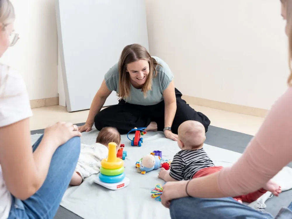 Kursleiterin sitzt lächelnd zwischen Müttern und Babys, die auf einer Decke mit Spielzeug spielen