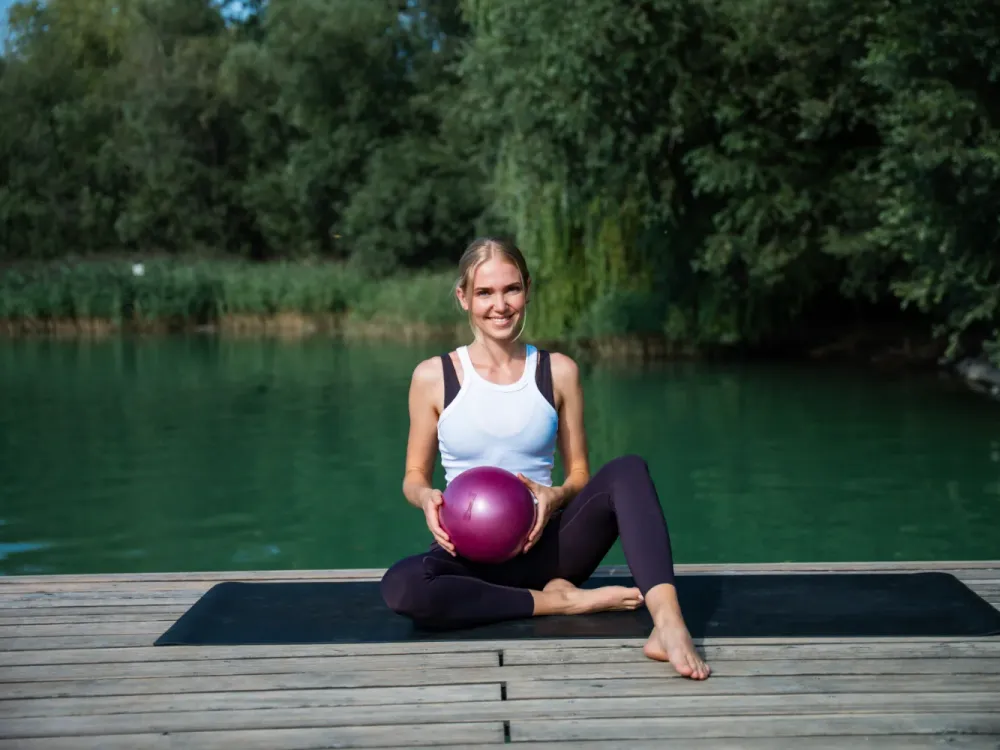 Frau sitzt lächelnd auf einem Steg am See und hält einen Pilatesball in den Händen
