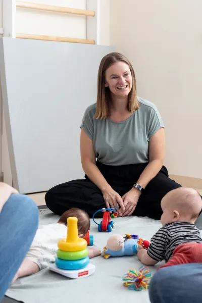 Kursleiterin sitzt lächelnd im Schneidersitz auf einer Matte zwischen zwei Müttern, während zwei Babys mit Spielzeug auf einer Decke liegen
